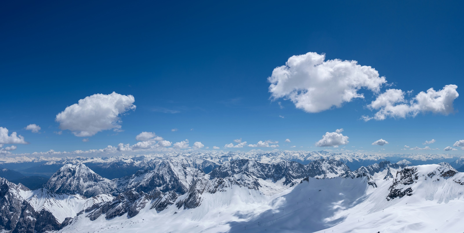 A panorama of snow covered mountains, blue sky and a few fluffy white clouds.