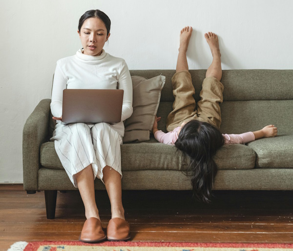 Mother with a laptop on a couch with daughter next to her on her back with feet up the wall.