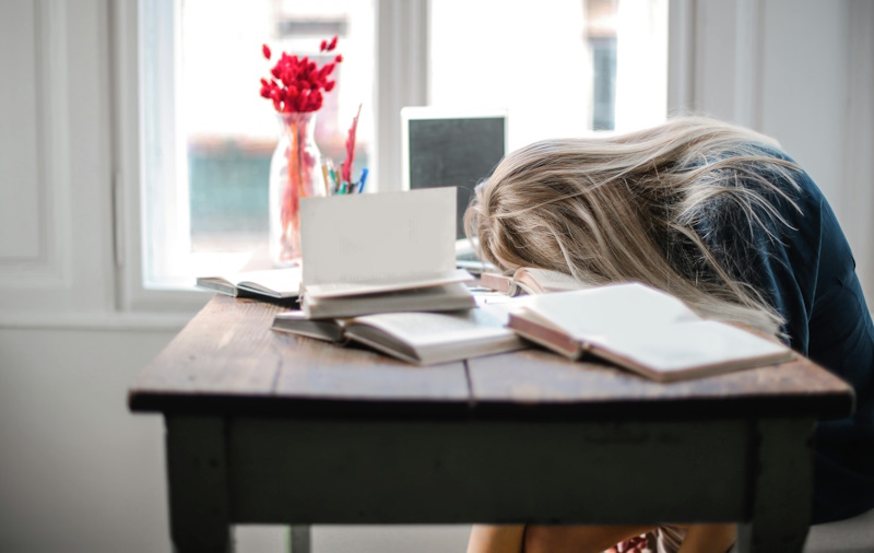 Woman with her forehead resting on open books at a desk.