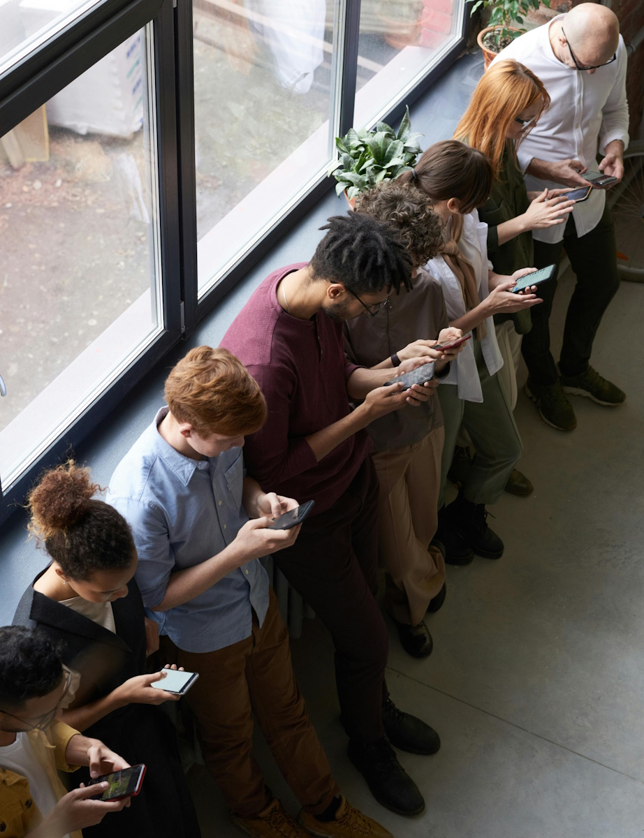 Top view of a line of people leaning against a window in a corridor, all looking at their mobile phone.