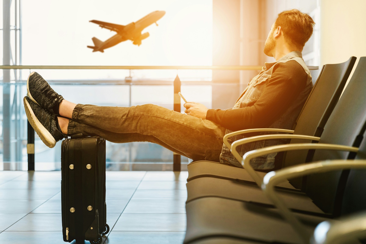 A man sitting at the airport looking out the window at a plane taking off.